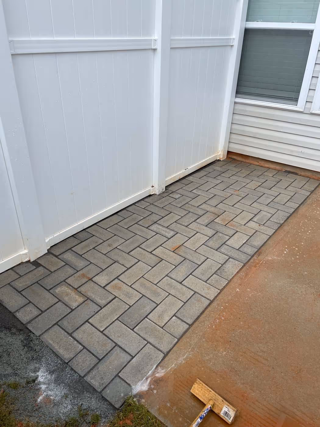 Newly installed gray brick paver patio adjacent to a white vinyl fence and house siding with a wooden tool resting on nearby dirt.