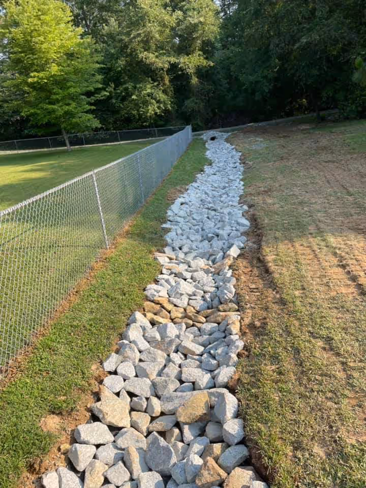 A dry creek bed lined with large gray and tan rocks runs alongside a chain-link fence on a grassy area with trees in the background.