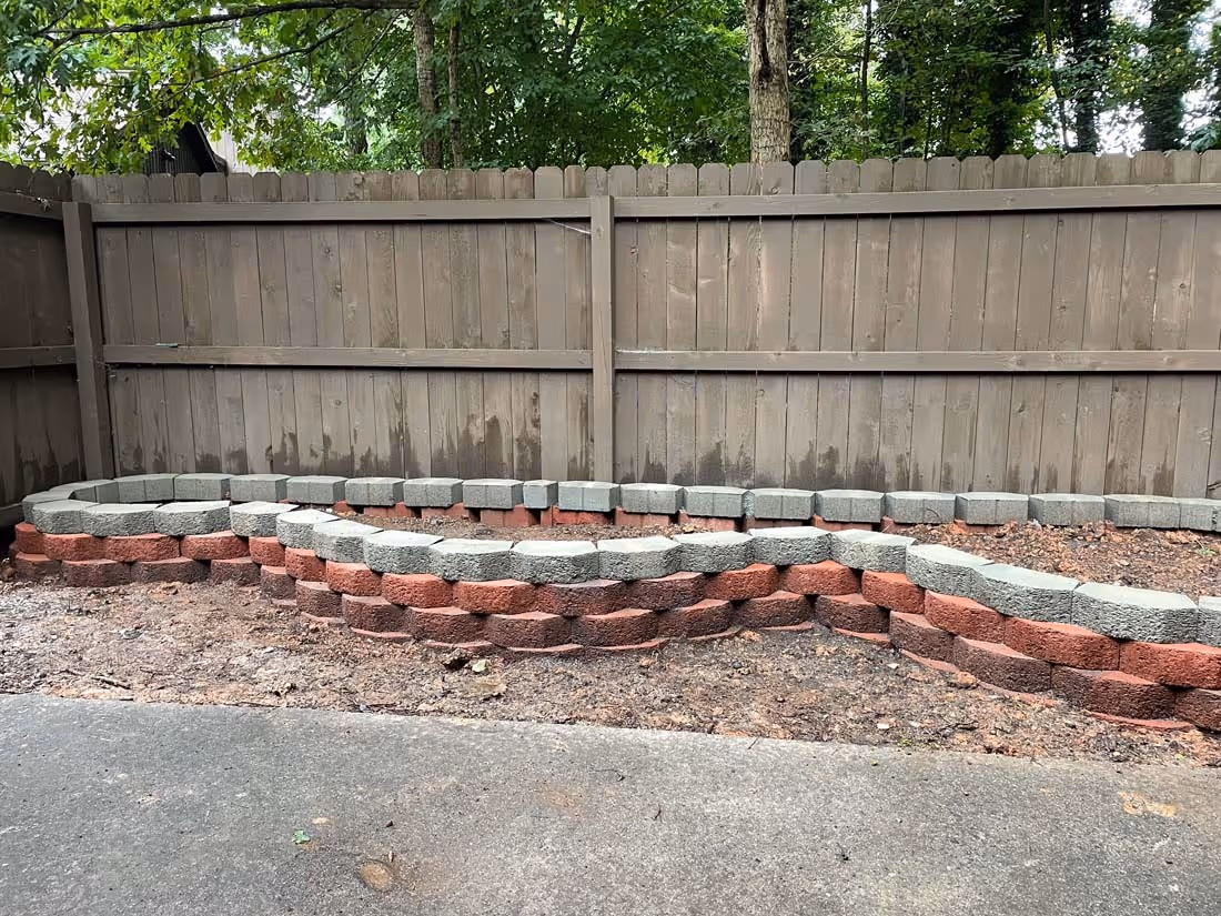 Two-tiered garden bed made of red and gray concrete bricks in front of a wooden fence with green trees behind.