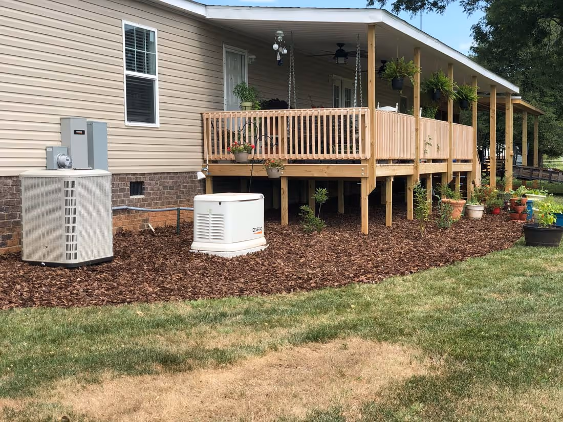 Side view of a house with beige siding, a raised wooden porch with hanging plants, and mulch-covered ground with installed air conditioning and generator units.