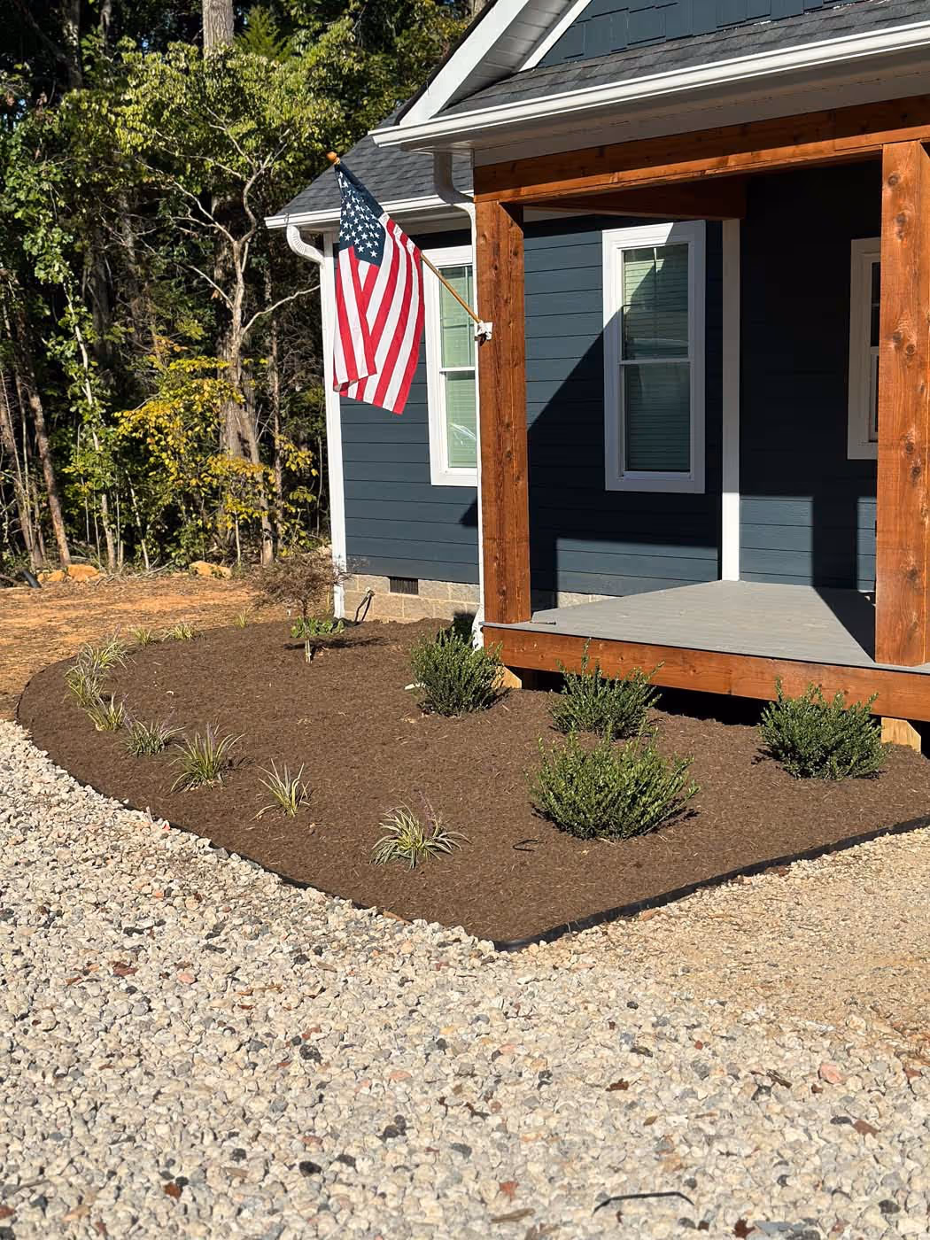 Front porch of a house with wooden pillars, blue siding, an American flag, and a mulched garden bed with small bushes and plants bordered by gravel.