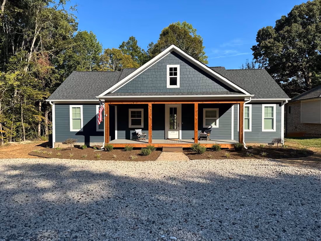 Single-story blue house with a front porch featuring wooden columns, two chairs, and an American flag, surrounded by trees and gravel driveway.