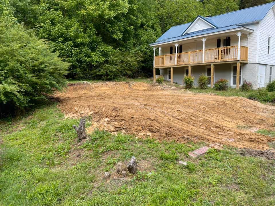 White elevated house with a blue metal roof and wooden porch overlooking a freshly cleared and leveled dirt area surrounded by green grass and trees.