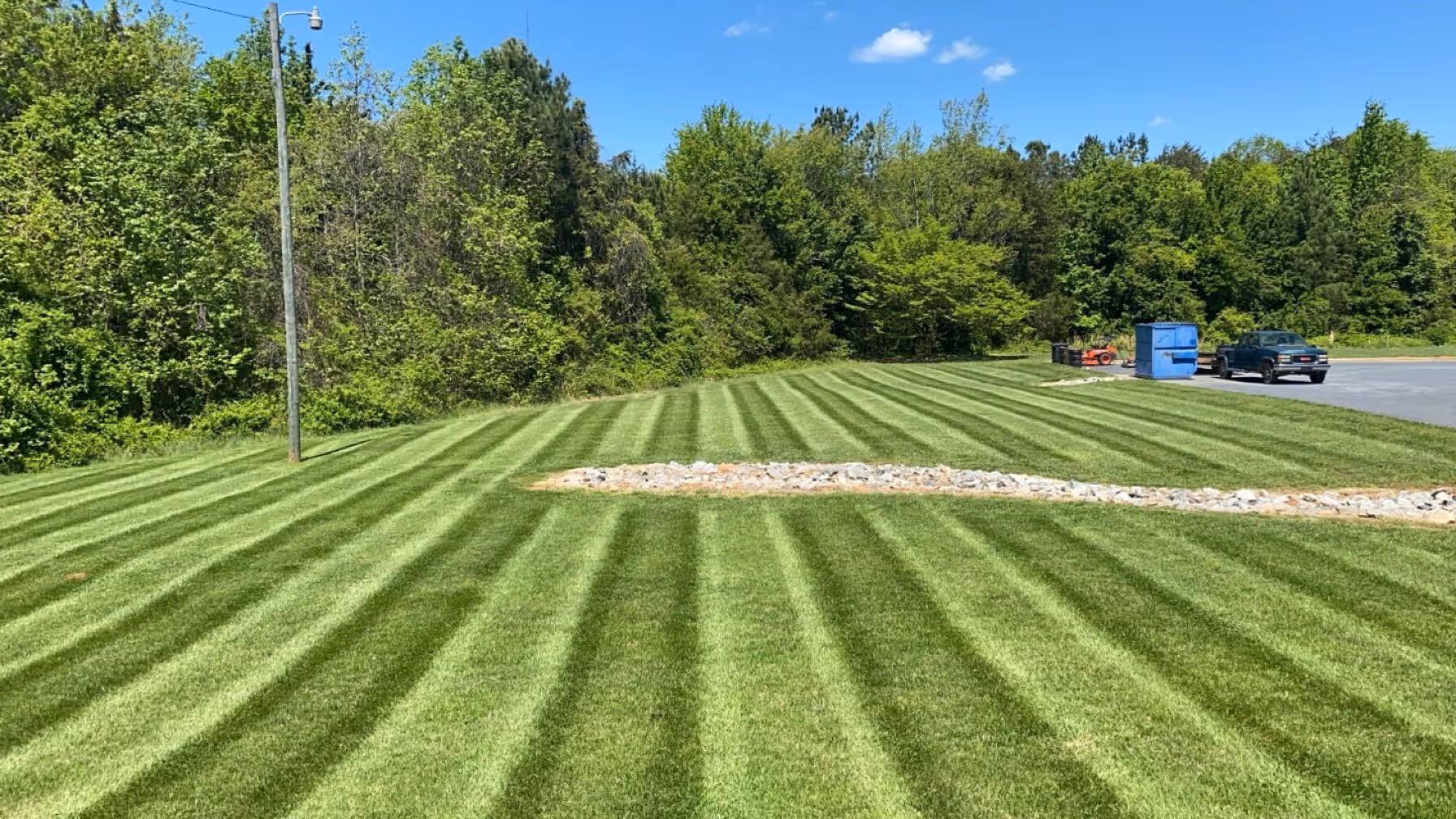 Well-maintained lawn with alternating light and dark green mowing stripes beside a wooded area under a clear blue sky.