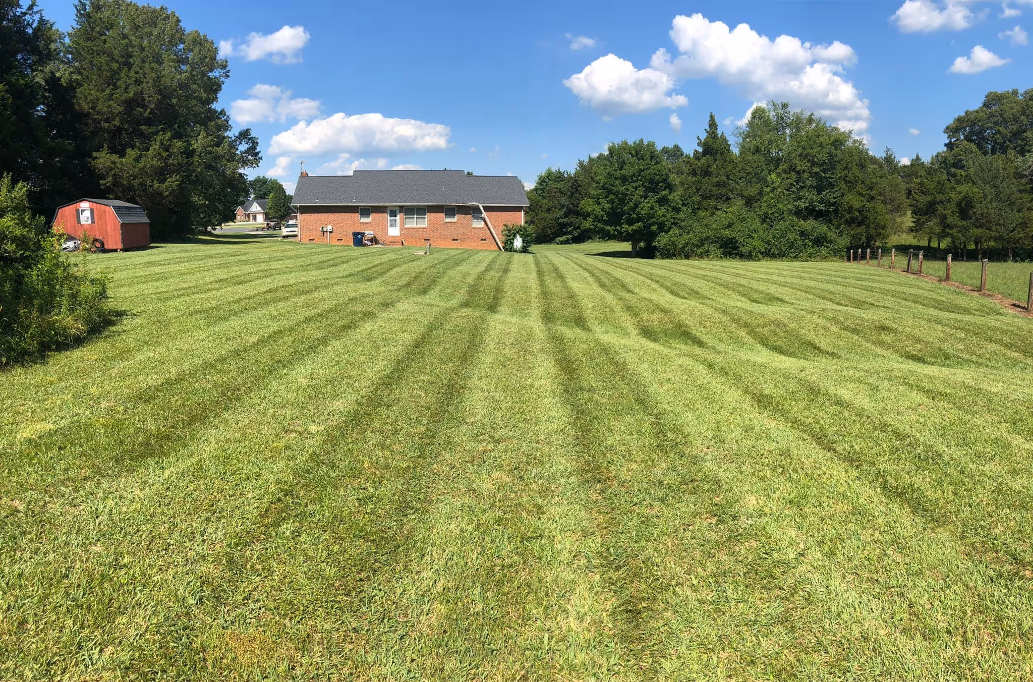 Large green lawn with visible mowing stripes in front of a brick house under a blue sky with scattered white clouds.