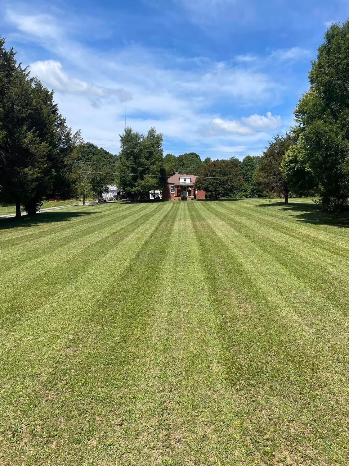 Wide, neatly mowed lawn with striped grass leading to a small brick house surrounded by trees under a partly cloudy blue sky.