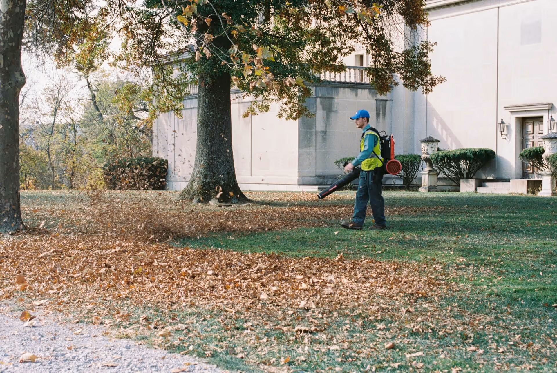 A worker in a blue cap and yellow vest using a backpack leaf blower to clear fallen leaves on a lawn near a large stone building.