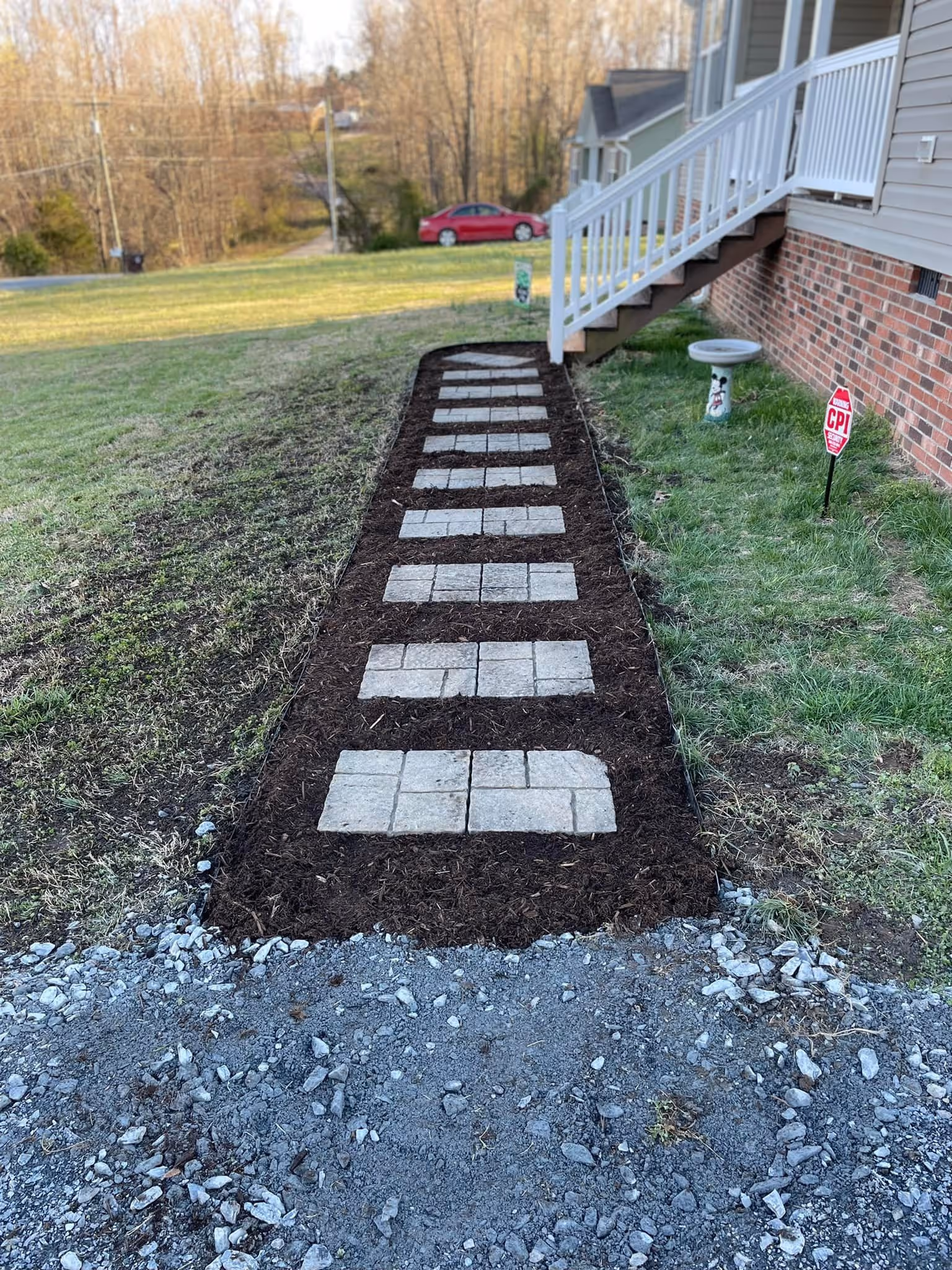 Mulched garden pathway with square stone pavers leading to house stairs beside a brick wall.