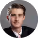 Portrait of a young man with short light brown hair wearing a black suit, white shirt, and pink tie against a gray background.